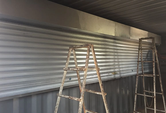 A storefront with a closed roller shutter covering the window and two ladders
